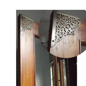 Puede incluir: Primer plano de un marco de puerta de madera con escuadras de esquina plateadas y ornamentadas. Las escuadras presentan un diseño floral detallado y arremolinado. La madera tiene un tono marrón cálido y las escuadras están fijadas con pequeños tornillos.