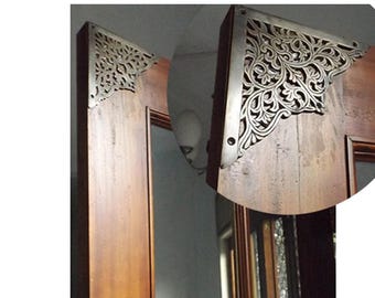 May include: Close-up of a wooden door frame with ornate, silver-colored corner brackets. The brackets feature a detailed, swirling floral design. The wood has a warm, brown tone, and the brackets are attached with small screws.
