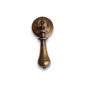 May include: A brass-toned metal drop pull handle featuring a circular backplate and a vertical, tapered pendant. The hardware has a weathered, antique finish with visible dark patina accents along the edges and the central stem structure.