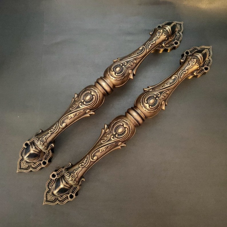 Pode incluir: Um par de ma&ccedil;anetas de porta douradas ornamentadas com intrincados desenhos florais. As ma&ccedil;anetas s&atilde;o feitas de metal e t&ecirc;m um acabamento polido.