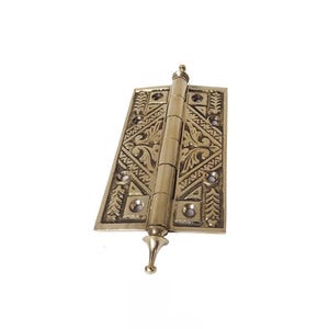May include: A rectangular brass hinge featuring an intricate embossed floral and geometric pattern across its surface. The hinge includes a central barrel with decorative finials at both the top and bottom ends, along with six pre-drilled screw holes.