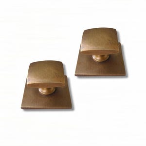 Op de afbeelding: Twee bijpassende kastknoppen van geborsteld messing. Elke knop heeft een vierkante basisplaat en een afgeronde, rechthoekige handgreep. Het metaal heeft een warme, gedempte goudtint met een gladde en consistente afwerking.