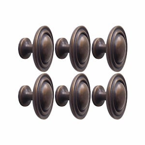 May include: A set of six round cabinet knobs with a dark bronze finish. Each knob features a central dome surrounded by a raised circular ring, creating a layered profile. The metal surface has a smooth, slightly weathered appearance.