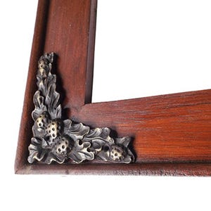 Pode incluir: Uma moldura de madeira para fotos com cantos de metal ornamentados com um design de folhas e bagas. A moldura é de cor marrom avermelhada escura.