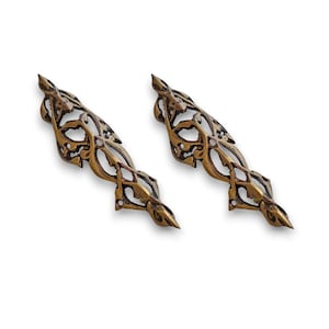 May include: Two ornate, antique-style brass decorative elements. Each element has a symmetrical, elongated shape with intricate openwork patterns and pointed ends. The brass has a warm, aged patina.