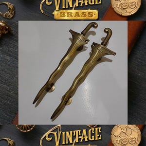Puede incluir: Dos manijas de puerta vintage de latón con un diseño ornamentado. Las manijas tienen una forma curva, similar a una espada, con empuñaduras texturizadas y elementos decorativos. Se muestran sobre un fondo neutro.