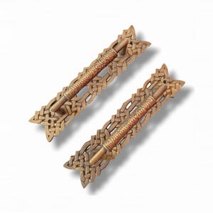 May include: Two metallic drawer pulls featuring a central cylindrical bar with a textured, weathered finish. Each handle is mounted on an ornate, rectangular backplate displaying an intricate, interwoven lattice pattern in a bronze tone.
