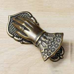 Könnte beinhalten: Ein messingfarbener Metallclip in Form einer skulptierten Hand mit einem Ring am Finger. Die Basis und der Ärmelaufschlag sind mit komplexen Prägemustern verziert, darunter kleine Fische und geometrische Formen in metallischer Optik.