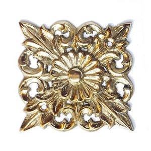 May include: A gold-toned metal decorative ornament with a floral design. The ornament features a central flower with intricate details and a raised center.
