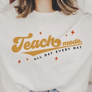 Könnte beinhalten: Weißes Sweatshirt mit dem Text "Teach mode all day every day" in einer Retro-Schriftart in Gelb-Gold. Der Text ist diagonal mit einer Baseball-Linie darunter. Es gibt kleine rote Sterne, die um den Text verstreut sind.