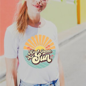 May include: White t-shirt with a retro-style graphic design featuring a sun, palm trees, and the text "Here Comes the Sun".