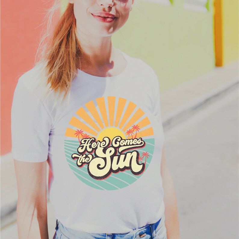 May include: White t-shirt with a retro-style graphic design featuring a sun, palm trees, and the text "Here Comes the Sun".