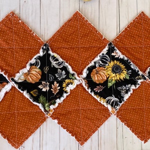 May include: A quilted table runner featuring a repeating pattern of alternating squares. The squares are a rust-colored fabric with white polka dots and black fabric with pumpkin and sunflower designs. The edges have a ruffled trim.