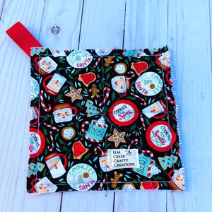 May include: A square, quilted Christmas-themed pot holder with a black background. The fabric features Santa Claus, gingerbread men, cookies, candy canes, and the text "Cookies for Santa." A red ribbon loop is attached to the corner.