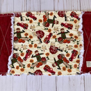 Op de afbeelding: Een rechthoekige, crèmekleurige rag quilt met een herfst oogstpatroon. Het patroon bevat vogelverschrikkers, pompoenen, zonnebloemen, rode vrachtwagens, schuren en schapen. De quilt heeft een bordeauxrode rand en een label met "Elm Creek Crafty Creations."