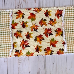 May include: A rectangular table runner with a patchwork design. The runner features alternating sections of gold floral fabric, green and brown plaid, and a centre panel with autumn leaves. The edges have a ruffled trim.