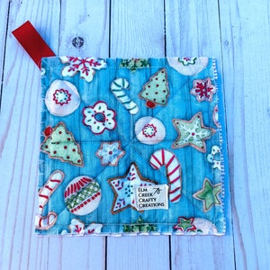 Puede incluir: Un salvamanteles navideño cuadrado y acolchado, con un fondo azul claro y varios diseños de galletas. Las galletas incluyen hombres de jengibre, bastones de caramelo y estrellas. Una cinta roja está unida a la esquina. La etiqueta dice "Elm Creek Crafty Creations."
