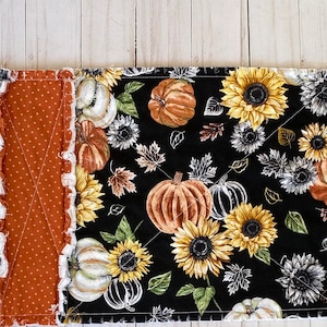 May include: A long, rectangular table runner with a fall theme. The runner features alternating panels of black fabric with sunflowers and pumpkins, and orange fabric with white polka dots. White ruffled edges add texture.