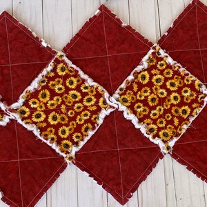 Peut inclure: Chemin de table fait à la main avec un motif de losanges répété. Des losanges en tissu bordeaux alternent avec des losanges à imprimé tournesol. Les bords sont blancs et effilochés, ajoutant de la texture.
