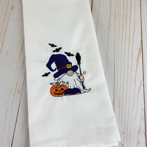 May include: White Halloween-themed hand towel with an embroidered gnome wearing a witch hat and holding a broom. A carved orange pumpkin and black bats are also embroidered on the towel. The towel is folded and has a textured appearance.