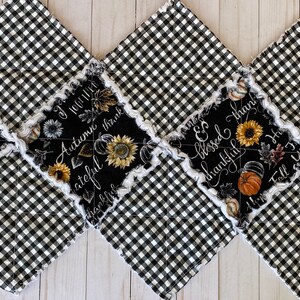 May include: A quilted table runner with a black and white checkered pattern. The center panels feature black fabric with fall-themed text, sunflowers, and pumpkins. The edges have a white, frayed trim.