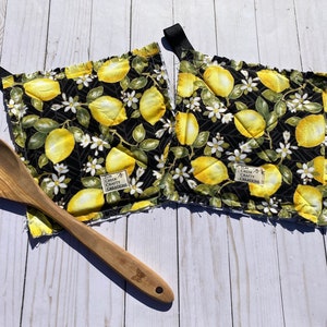 May include: Two square pot holders with a black background, featuring a lemon and floral pattern. Each pot holder has a black loop for hanging. A wooden spoon rests on the surface.