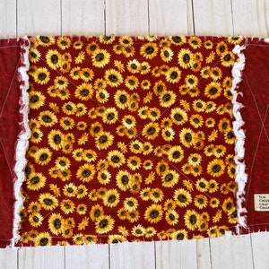 May include: A rectangular, handmade, rag quilt featuring a sunflower pattern on a burgundy background. The quilt has burgundy borders and white, fringed edges. The quilt is labeled "Tlm Creek Crafty Creations".