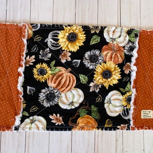 Pode incluir: Toalha de mesa acolchoada retangular com tema de outono. O centro apresenta um fundo preto com girassóis, abóboras e folhas. As bordas são bordadas com um tecido cor de ferrugem com bolinhas brancas e uma borda branca com babados. A toalha de mesa tem uma etiqueta que diz "ELM CREEK CRAFTY CREATIONS."