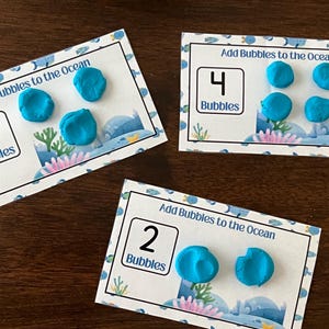 Puede incluir: Tres tarjetas educativas rectangulares tituladas Add Bubbles to the Ocean, cada una con un número y bolas de arcilla azul correspondientes. Las tarjetas muestran los números 2, 3 y 4, con cantidades coincidentes de pequeñas burbujas de arcilla azul.