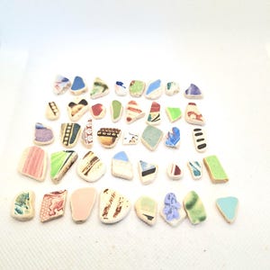 May include: A collection of forty-two small, irregular ceramic shards featuring a variety of colours including blue, green, pink, red, brown, and black. Each piece displays unique patterns, stripes, or solid glazes on a white ceramic base.