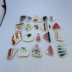 May include: A collection of 20 small, white ceramic tiles with various colourful patterns. The tiles are arranged in rows on a white surface. The patterns include geometric shapes, floral designs, and abstract art.