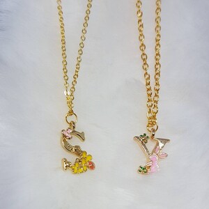Puede incluir: Dos collares de oro con dijes de letras. El dije 'S' tiene flores amarillas y una mariposa rosa. El dije 'Y' tiene un conejito rosa y hojas verdes.