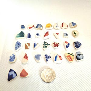 May include: A collection of thirty-one small, smooth, multi-coloured ceramic sea glass shards featuring various patterns and shapes, arranged in rows alongside a single silver-coloured coin for scale, showcasing diverse blue, red, and white decorative designs.