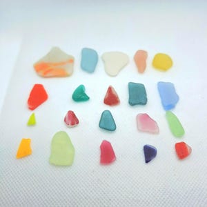 Könnte beinhalten: Eine Sammlung farbenfroher Seeglasstücke in verschiedenen Formen und Größen. Die Stücke sind in den Farbtönen Rot, Orange, Gelb, Grün, Blau und Lila gehalten. Das Seeglas ist auf einer weißen Oberfläche angeordnet.