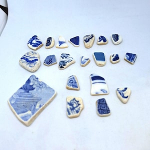 May include: A collection of blue and white ceramic shards with various patterns and designs. The fragments vary in size and shape, with some featuring floral or geometric motifs. The pieces are arranged on a white surface, showcasing their aged appearance.