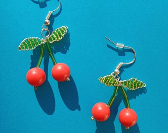 May include: A pair of cherry earrings with green leaves and red beads. The earrings are on a blue background.