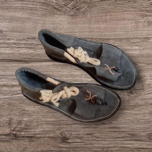 Könnte beinhalten: Ein Paar graue Wildleder-Sandalen mit cremefarbenen Seildetails. Die Sandalen haben einen Zehensteg im Zehensteg-Stil und eine genähte Sohle. Das Schuhwerk wird auf einer Holzoberfläche präsentiert.