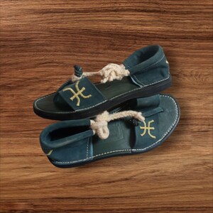 Könnte beinhalten: Ein Paar türkisfarbene Wildledersandalen mit einem Seilriemen und einem goldgestickten Symbol. Die Sandalen haben eine dunkle Sohle und werden auf einer Holzoberfläche präsentiert. Einzigartige Schuhe.