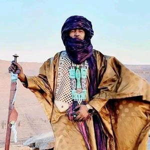 May include: A person wearing traditional desert attire, including a turban, scarf, and flowing robe in shades of gold and purple. They hold a sword with a wooden handle. The background shows a desert landscape.