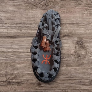 Könnte beinhalten: Graue Wildledersandale mit geflochtenen Riemen und einer braunen Quaste. Die Sandale hat ein orangefarbenes, gesticktes Symbol. Die Sohle ist schwarz. Die Sandale wird auf einer Holzoberfläche präsentiert.