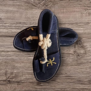 Könnte beinhalten: Ein Paar marineblaue Wildleder-Sandalen mit cremefarbenen Seildetails und gelben Nähten. Die Sandalen haben ein Zehenschlaufendesign und einen kleinen Absatz. Sie werden auf einer Holzoberfläche präsentiert.