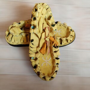 Könnte beinhalten: Ein Paar gelbe Ledersandalen mit aufwendigen Ausschnitten. Die Sandalen haben ein gewebtes Muster auf dem Obermaterial und eine schwarze Sohle. Dekorative Quasten und gestickte Symbole ergänzen das Schuhwerk.