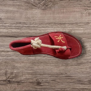 Könnte beinhalten: Eine rote Wildledersandale mit cremefarbenen Seildetails und einem goldgestickten Symbol. Die Sandale hat einen Zehenriemen und ein geschlossenes Zehendesign. Die Sohle ist mit weißem Faden genäht.