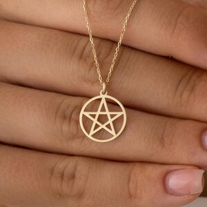 Puede incluir: Un collar con colgante dorado con un diseño de pentagrama dentro de un marco circular. El collar se muestra en una cadena delicada. El colgante lo sostiene una persona.