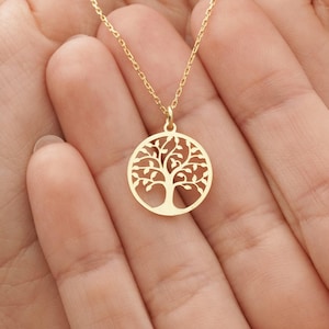 Puede incluir: Un collar dorado con un colgante circular con un diseño de árbol de la vida. El colgante está suspendido de una delicada cadena. El collar se sostiene en manos abiertas sobre un fondo neutro.