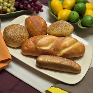 May include: A collection of assorted baked bread items on a rectangular off-white platter, including a round seeded roll, a smooth oval loaf, a textured seeded bun, a long golden-brown loaf, and a single elongated, dark-crusted bread stick.
