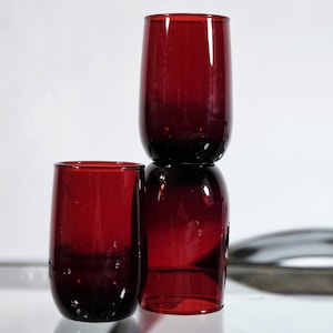 May include: Two cylindrical drinking glasses made of deep red translucent glass. One glass stands upright on the left, while the other is positioned vertically on top of a third inverted glass, creating a stacked arrangement of three tumblers.