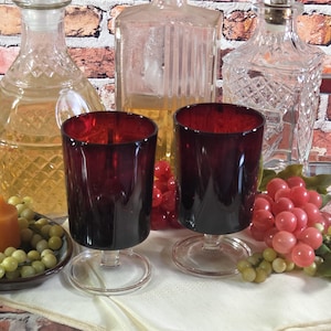 May include: Two matching deep red glass goblets with smooth cylindrical bowls and clear glass stems and circular bases. The glasses are positioned in front of three clear glass decanters and decorative clusters of artificial grapes.