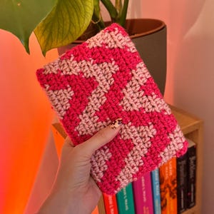 May include: A hand-crocheted book cover featuring a striking zigzag design in shades of pink. The cover is held up, revealing a bookshelf with books. The crochet is tightly worked, creating a textured finish.