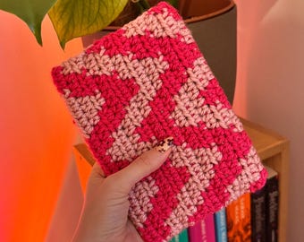 Puede incluir: Una funda de libro hecha a mano con un llamativo diseño en zigzag en tonos rosa y rosa claro. La funda se sostiene frente a una estantería con lomos de libros coloridos. El ganchillo es apretado.